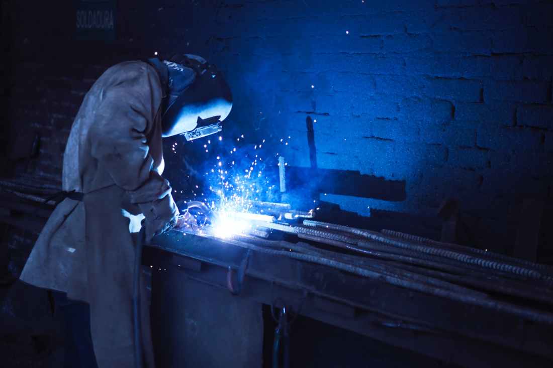 man wearing welding helmet welding metal near gray brick wall